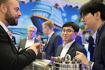 Mehrere Personen stehen an einem Messestand und betrachten gemeinsam ein technisches Metallbauteil. Im Hintergrund ist eine großformatige Darstellung eines Schiffes zu sehen.