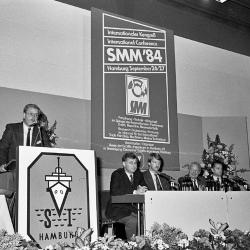 Ein Schwarz-Weiß-Bild von einer früheren Messe, auf dem ein Redner von der Bühne spricht; das Bild wurde aus der vordersten Reihe aufgenommen