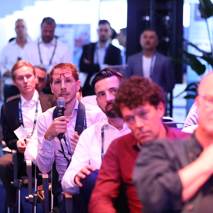 Eine Person sitzt bei einer Veranstaltung im Publikum und hält ein Mikrofon, während eine Frage gestellt oder ein Beitrag gemacht wird. Die Umgebung ist ein gut besuchter Konferenz- oder Networking-Bereich mit mehreren sitzenden und stehenden Teilnehmenden im Hintergrund, die aufmerksam zuhören.
