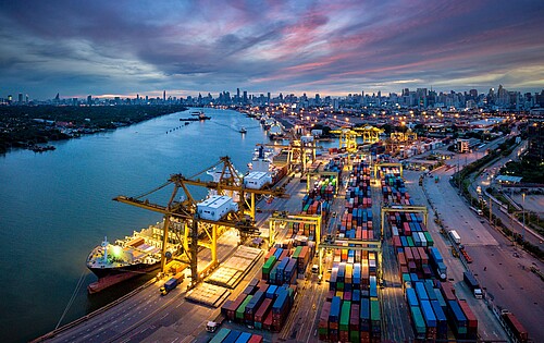Luftaufnahme Hafen mit Kran, der Container auf Schiff lädt, vor Kulisse nächtlicher Skyline von Bangkok.