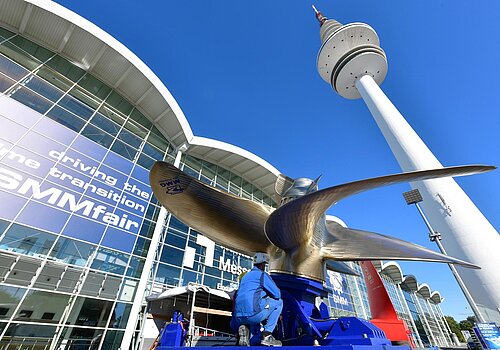 Ein goldenes Windrad, welches vor dem Hamburg Messe Gebäude installiert wird. Das Messegebäude wird schräg von unten gezeigt. 