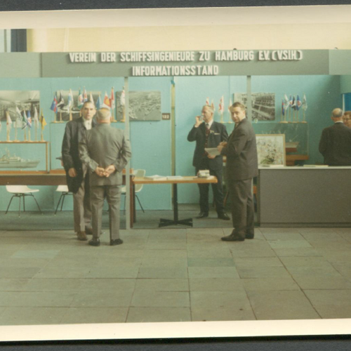 Historisches Bild des Informationsstandes des Vereins der Schiffsingenieure zu Hamburg E.V.. Mit zwei Tischen und mehreren Bildern an der Wand hängend.