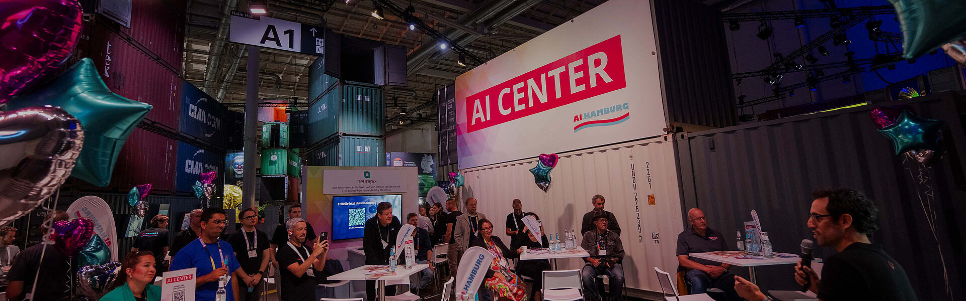 Ein seitlich aufgenommenes Bild in der AI Center Area, wo jemand ins Mikrofon spricht. Zuschauer sitzen an Tischen, während eine Person filmt