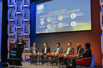 Podiumsdiskussion auf einer Konferenzbühne. Eine Person steht an einem Rednerpult mit der Aufschrift „Open Stage“, während sechs weitere Personen auf orangefarbenen Sesseln sitzen. Im Hintergrund zeigt eine große Leinwand Informationen zu einer maritimen Fachveranstaltung in Indien. Die Bühne ist in Blau- und Orangetönen beleuchtet und von dekorativen Wandpaneelen eingerahmt.