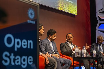 Mehrere Personen sitzen auf orangefarbenen Sesseln auf einer Bühne und nehmen an einer Podiumsdiskussion teil. Im Vordergrund steht ein Schild mit der Aufschrift „Open Stage“. Auf einem kleinen Tisch vor den Teilnehmenden stehen Wasserflaschen und Gläser. Der Hintergrund zeigt einen Teil der projizierten Präsentation sowie eine farbig beleuchtete Bühnenkulisse.