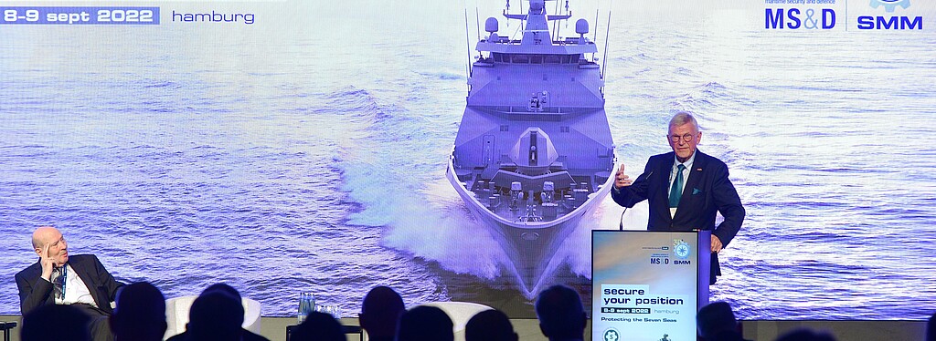 Ein Mann präsentiert etwas vor einem Publikum auf einer Leinwand die Wasser und ein Schiff abbildet. Außerdem steht darauf: Secure your Position und Daten.