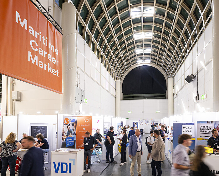 Eine Ausstellungshalle mit sichtbaren Ständen, Ausstellenden und Besuchenden. Im Vordergrund und Hintergrund ist der Hauptgang der Halle erkennbar, der die Stände miteinander verbindet.