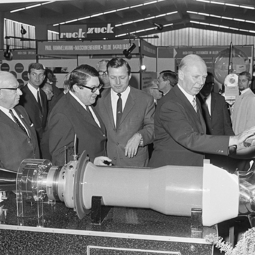 Historisches Bild einer Gruppe von Messebesuchern, die ein ausgestelltes Maschinenteil anschauen. Im Hintergrund sind weitere Messestände und -Besucher zu sehen.